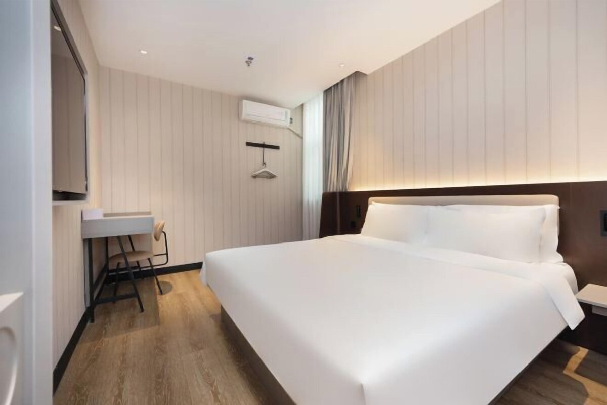 A modern hotel room with a large bed, a bedside table, a desk with a chair, and an air conditioning unit on the wall.