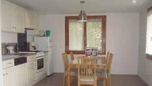 Cocina pequeña con armarios blancos, una estufa y un refrigerador. Hay una mesa de comedor con sillas y una ventana con cortinas decorativas.