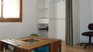 Una habitación con una mesa de madera, un radiador, un armario con ropa colgada y una silla.