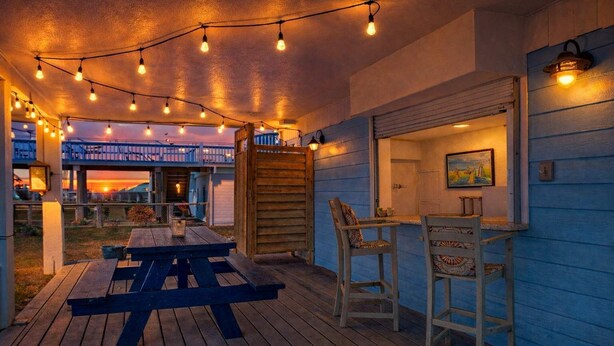 Un patio al aire libre con mesa y sillas de madera, luces colgantes y una vista de un edificio al fondo.