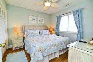 Un dormitorio con cama, dos lámparas de noche, ventilador de techo, ventana con cortinas y una mesita con un control remoto.