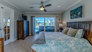 A bedroom with a bed, a ceiling fan, a nightstand, a painting, and a view of a pool through a sliding glass door.