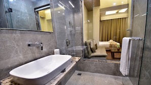A modern bathroom with a large bathtub, a glass shower enclosure, and a visible bedroom with a bed and curtains. - HOTEL CENTELLA  (Gwalior)