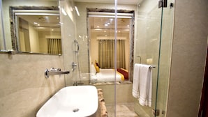 A modern bathroom with a large white sink, glass shower enclosure, and a visible bedroom with a bed and curtains in the background. - HOTEL CENTELLA  (Gwalior)