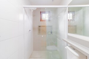 Un baño con azulejos blancos, cabina de ducha de vidrio y ventana sobre el lavamanos.