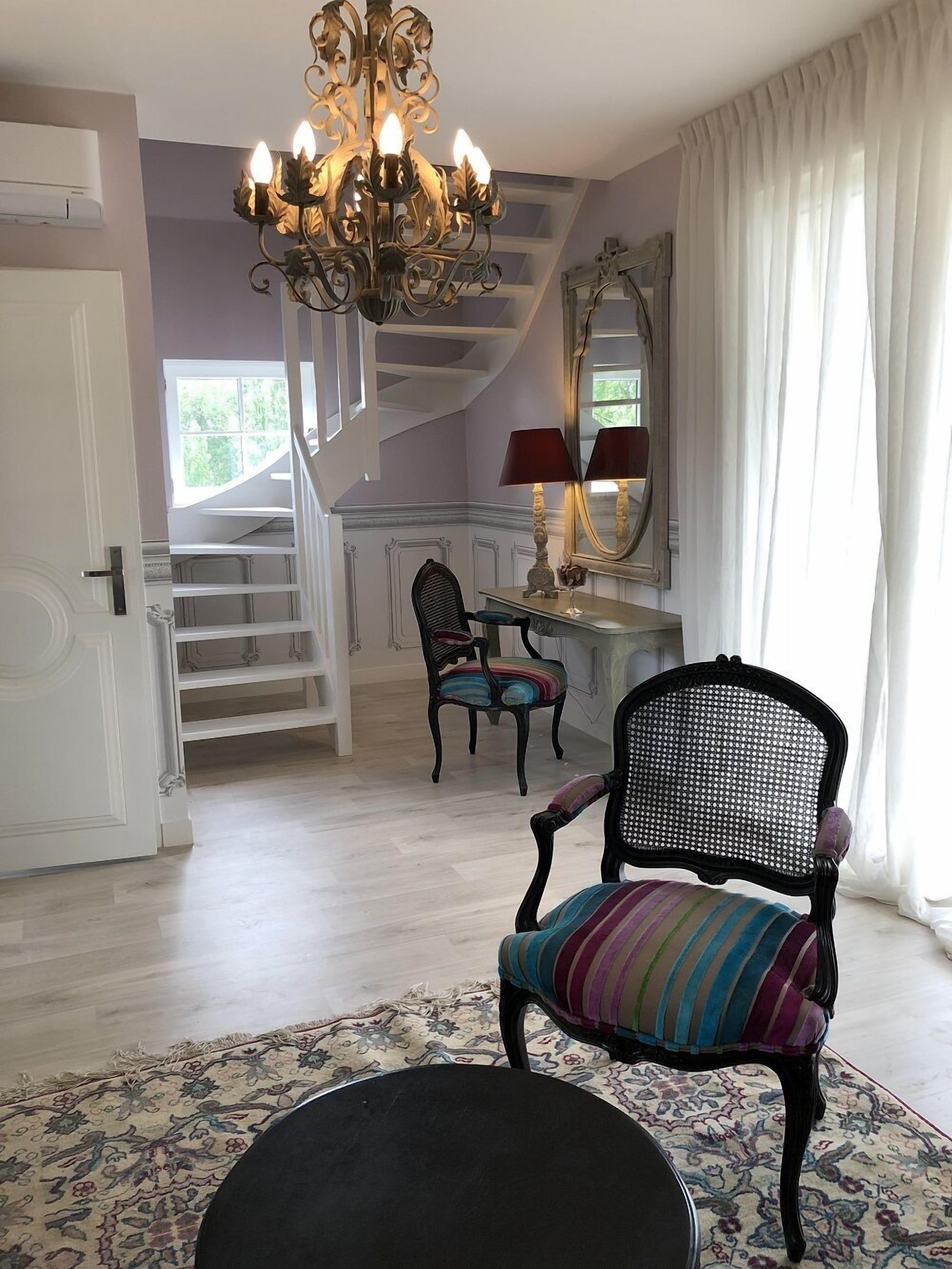 Una habitación con una araña, una silla con cojín a rayas, una mesa con lámpara y un espejo.