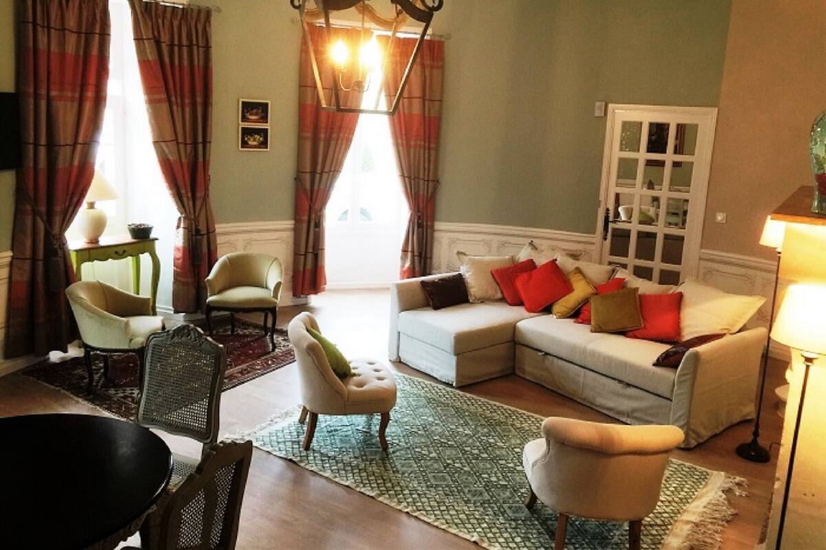 Un salon chaleureux avec un canapé d’angle, deux fauteuils, une table à manger et un lustre.
