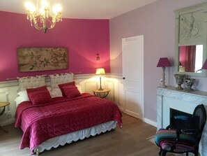 A bedroom with a bed, a chandelier, a fireplace, and a chair.