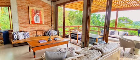 A spacious living area with a wooden ceiling, a large sofa, a dining table, and a view of greenery outside.