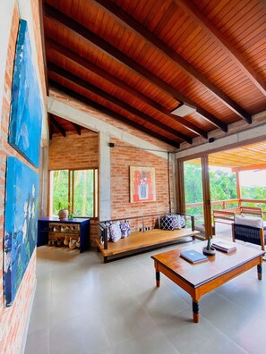 Una habitación con techo de madera, pared de ladrillo con un cuadro, banco de madera, una mesa y vista a follaje exterior.