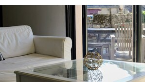 Een witte bank en een glazen tafel met een decoratieve vaas in een kamer met uitzicht op tuinmeubilair en een stenen muur.