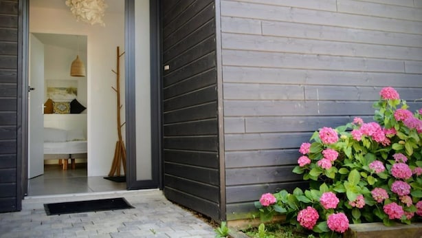 Een modern huis met een donker houten gevel, een zichtbaar interieur met een bed en een hanglamp, en een tuin met roze bloemen.