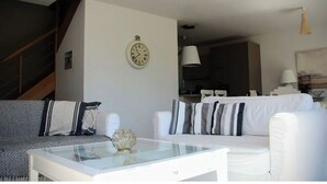 Een moderne woonkamer met een glazen salontafel, twee witte banken met gestreepte kussens, een klok aan de muur, en een trap die naar een ander niveau leidt.