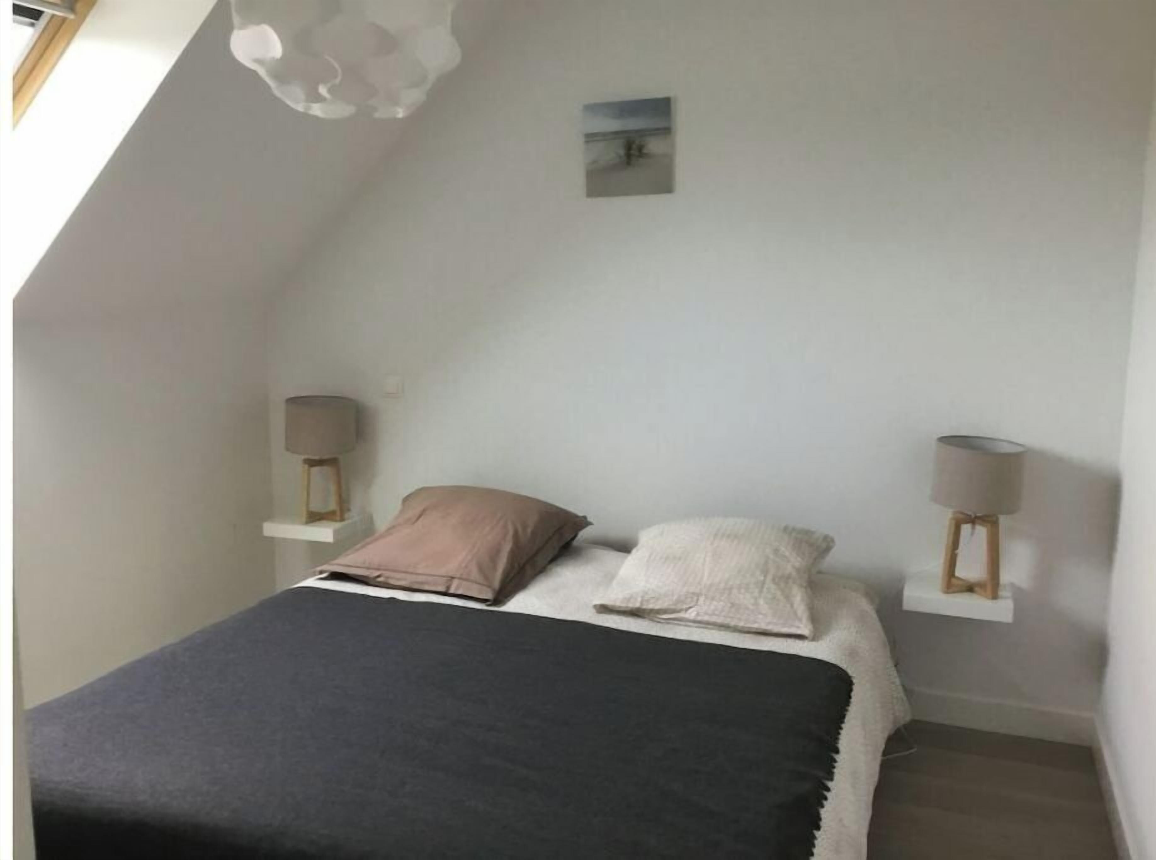 Ein Schlafzimmer mit schrägen Wänden, einem Bett mit zwei Kissen, einem Nachttisch mit Lampe und einem kleinen Bild an der Wand.