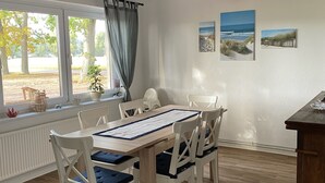 Ein Essbereich mit Holztisch und weißen Stühlen, ein Fenster mit Blick auf Bäume sowie zwei Wandbilder im Strandhausstil.
