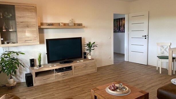 Ein Wohnzimmer mit Flachbildfernseher auf Holzfuß, Couchtisch mit Dekoration, Sofa und Essbereich mit einem Stuhl.