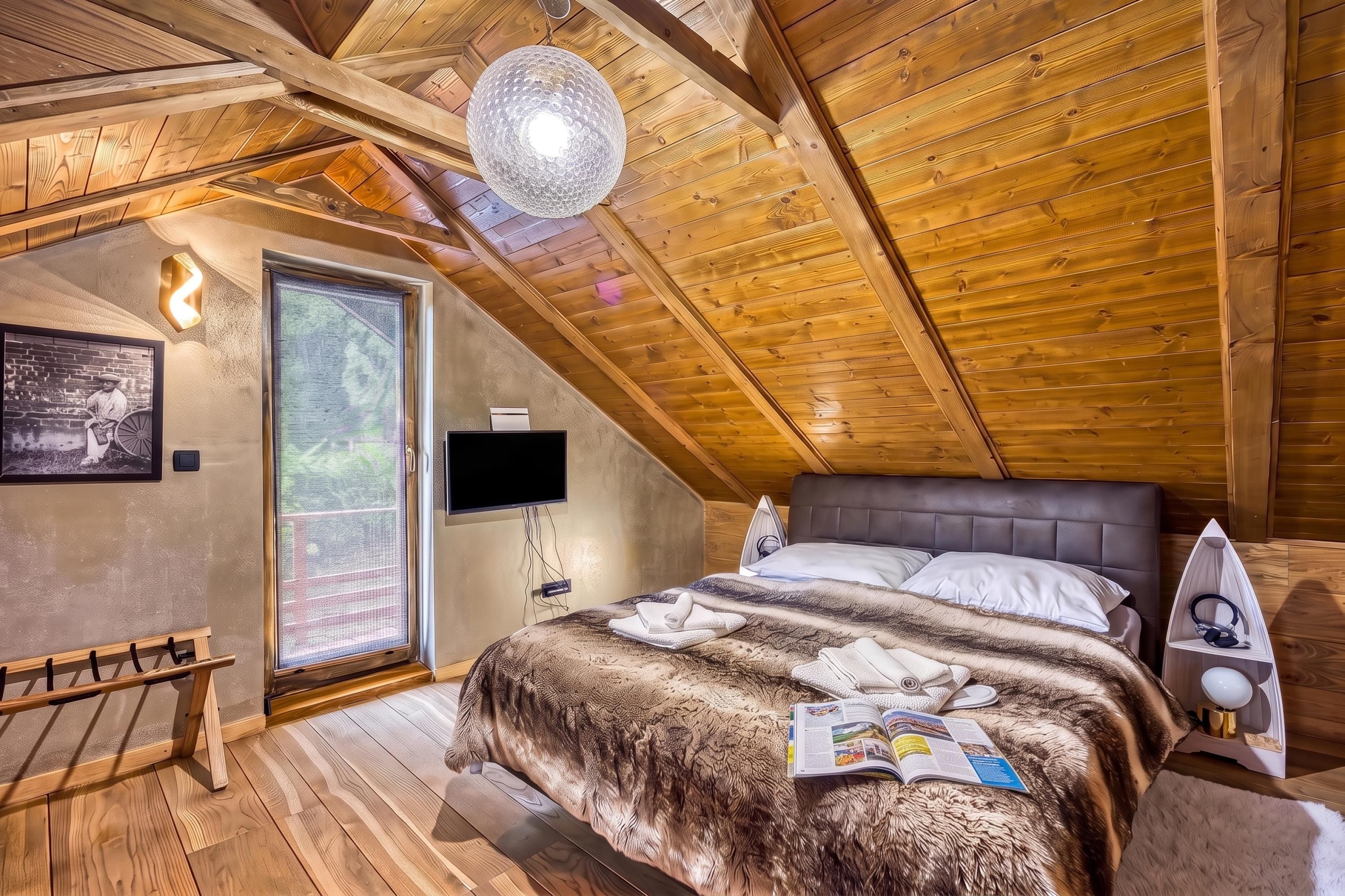 A cozy bedroom with a wooden ceiling, a large bed, a television, and a window.