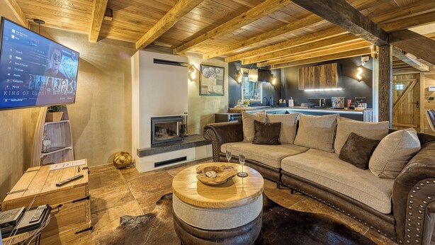 Ein gemütliches Wohnzimmer mit Holzbalkendecke, Kamin, einer Couch und einem Fernseher, der einen Film zeigt.