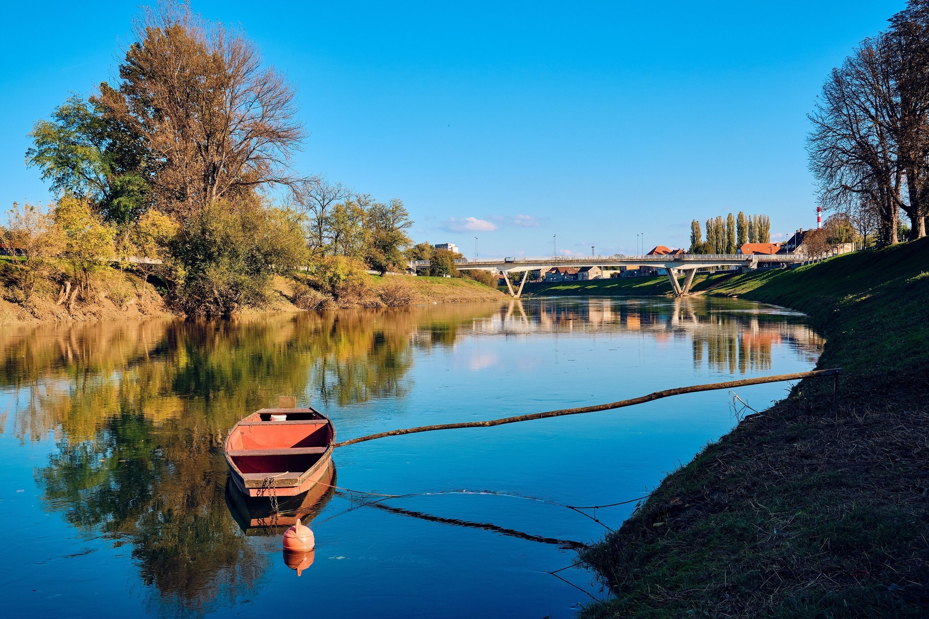 A calm river with a small boat docked on the left, a bridge in the background, and trees lining the riverbank.