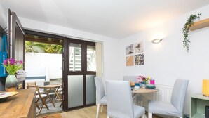 Un comedor con mesa y sillas de madera, una mesita auxiliar con un jarrón de flores y vistas a un espacio exterior con plantas a través de una puerta corredera de vidrio.