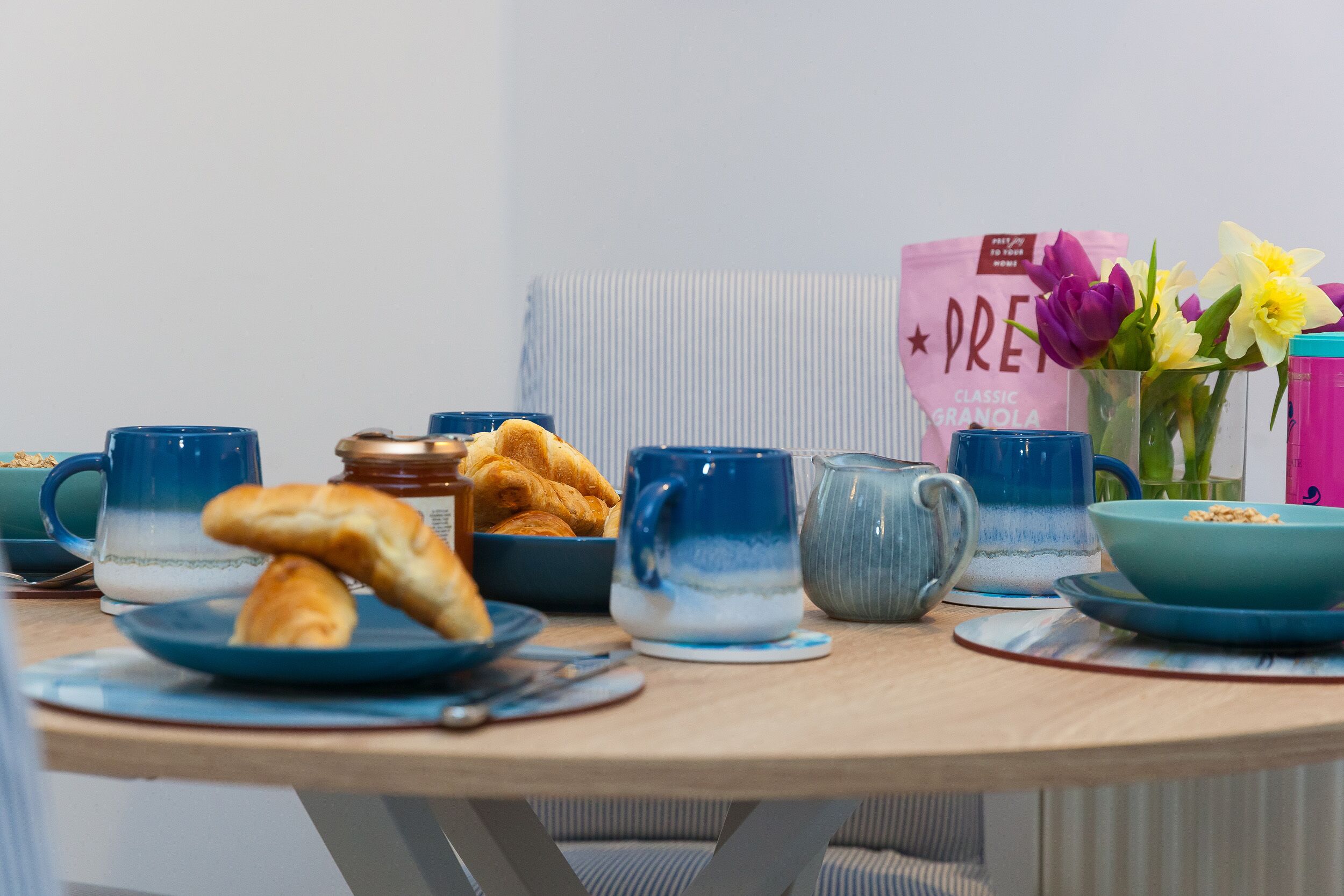 A breakfast table with croissants, a pink bag labeled "PREMIUM GRANOLA," and a vase with flowers.