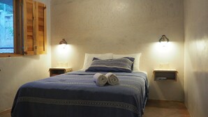 Una cama bien hecha con una colcha azul y dos almohadas, dos lámparas de noche y una ventana con contraventanas de madera.