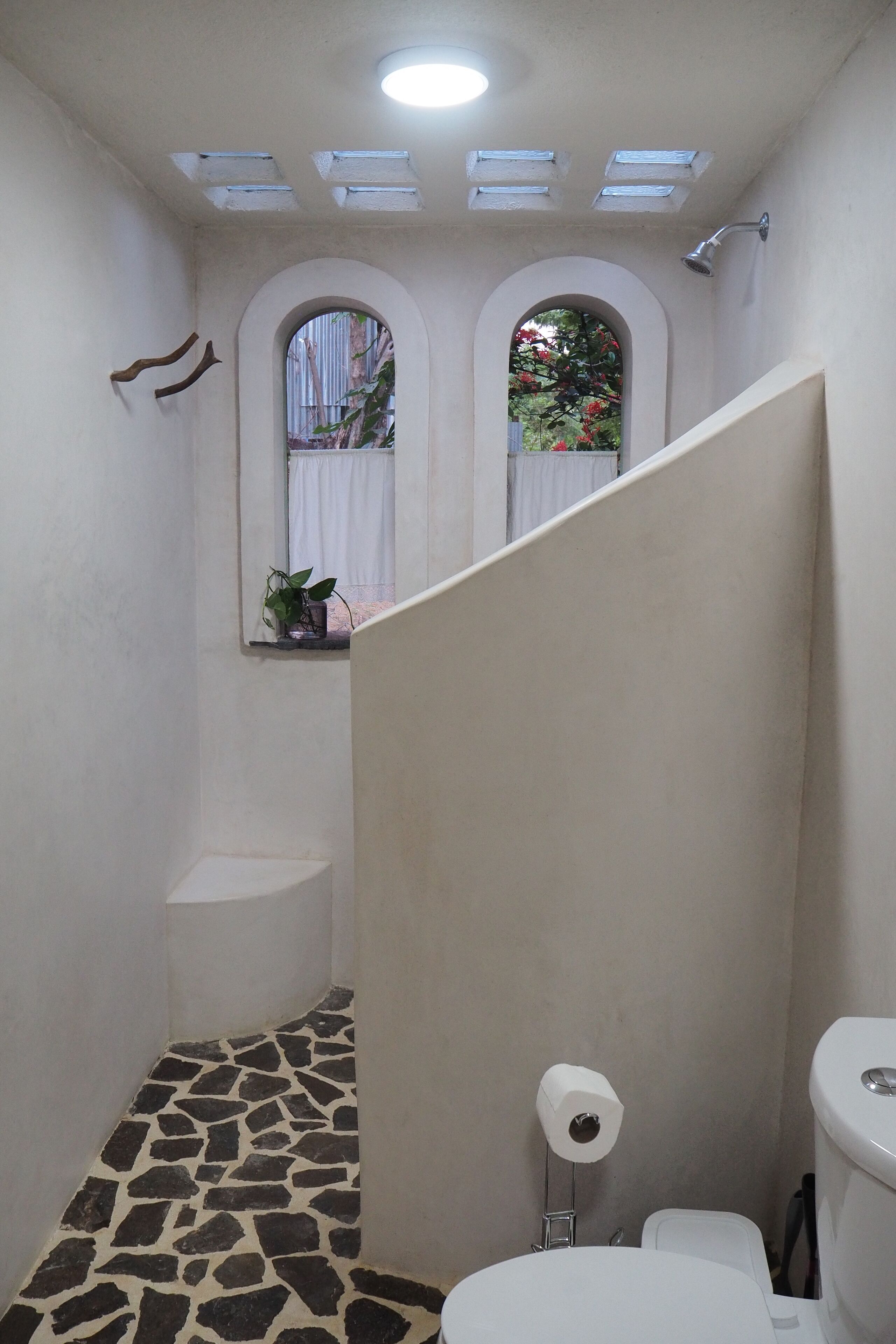 A bathroom with a stone-patterned floor, a toilet, a shower with a glass door, and a window with a view of greenery.