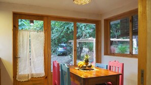 Una mesa de comedor de madera con sillas coloridas, una cesta tejida con frutas y vistas a un jardín a través de un ventanal.