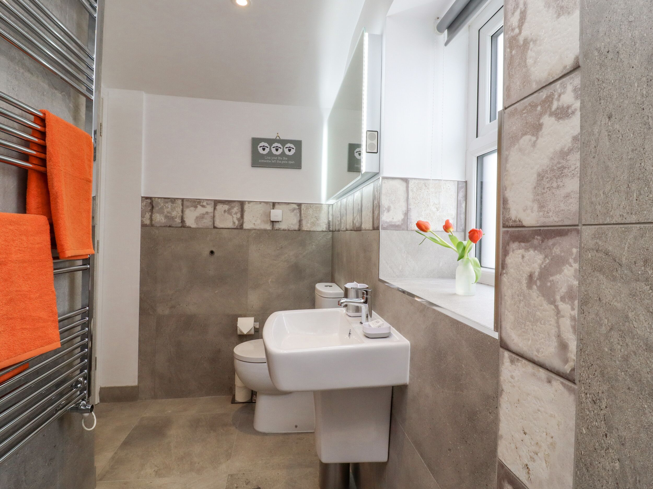 A bathroom with a white sink, a toilet, and a towel rack with orange towels. There is a window with a vase of flowers on the windowsill.
