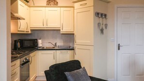 Una cocina compacta con armarios blancos, un microondas, estufa y lavaplatos. Un sillón negro con un cojín gris está en primer plano.