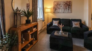 Una sala de estar con un sofá verde oscuro, una mesita de madera con una planta en maceta, una lámpara de pie y un cuadro en la pared.
