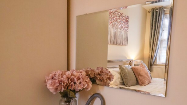 Un dormitorio con cama, mesita de noche, un jarrón con flores, un espejo y un adorno de pared.