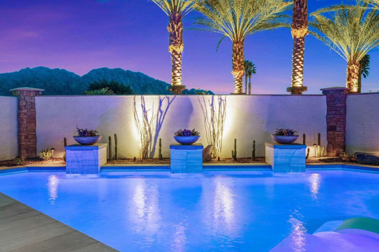 An outdoor swimming pool with a view of mountains at dusk, surrounded by palm trees and decorative planters.