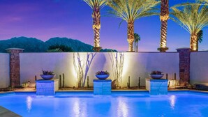 Une piscine extérieure offrant une vue sur les montagnes au crépuscule, entourée de palmiers et de jardinières décoratives.