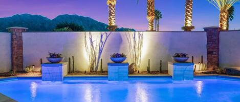 An outdoor swimming pool with a view of mountains at dusk, surrounded by palm trees and decorative planters.
