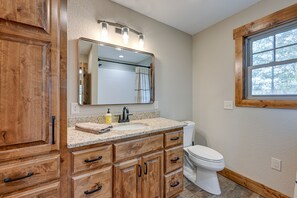 A bathroom with wooden cabinets, a sink with a mirror above it, a toilet, and a window. - Smoky Mountain Cabin Rental: Game Room, Fire Pit! (Andrews)