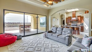A spacious living area with a grey sofa, red bean bag, and patterned rug. A large sliding glass door leads to an outdoor patio with a table and chairs. The kitchen is visible in the background with wooden cabinets and a range hood. - Unique Las Cruces Home w/ Patio + Gas Grill! (Las Cruces)
