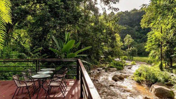 A wooden deck with outdoor seating overlooking a flowing river, surrounded by dense greenery and trees. - Zen Maekampong Waterfall Ryokan (.)