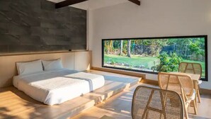 A modern bedroom with a large bed, a dining area with wicker chairs, and a view of a garden through a large window. - Zen Maekampong Waterfall Ryokan (.)