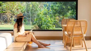 A person sitting by a large window with a view of greenery, a wicker chair, and a white sofa. - Zen Maekampong Waterfall Ryokan (.)