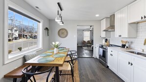 A modern kitchen with white cabinets, a wooden dining table, and a view of the street through a large window. - Cozy 4Bd Bungalow w/ Private Backyard Lounge and Game Room Mins to Downtown (Raleigh)