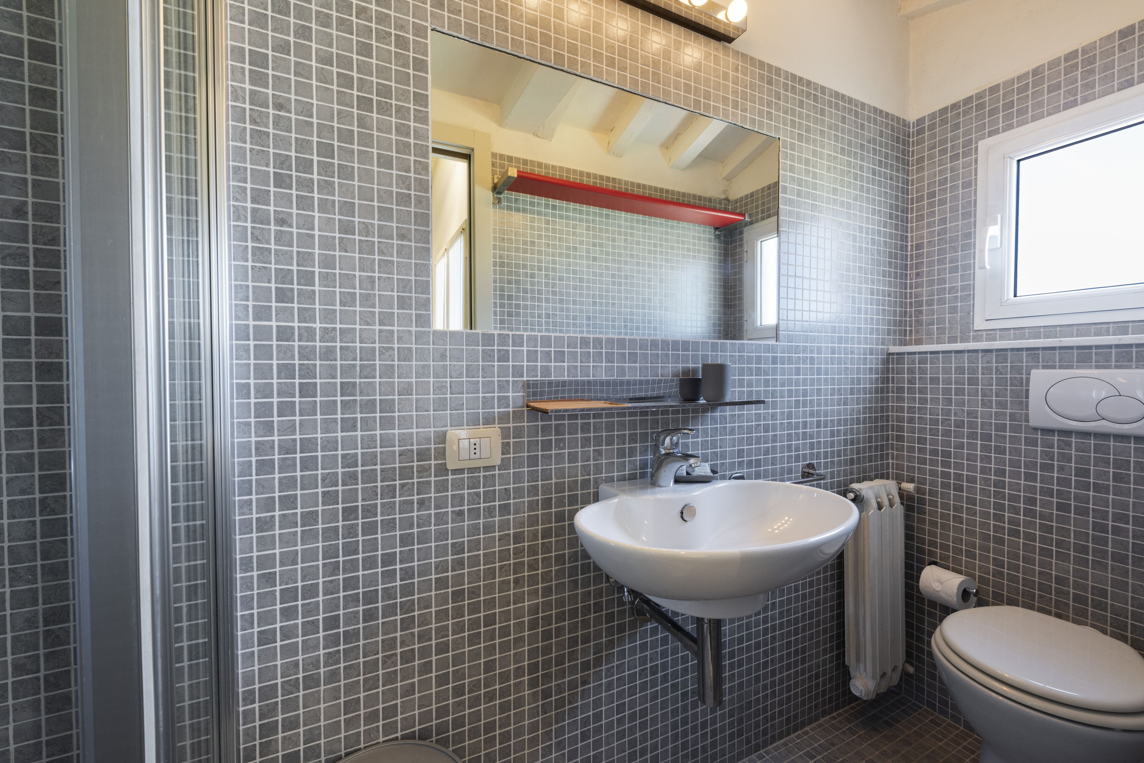 A bathroom with a tiled wall, a sink, a mirror, a toilet, and a window.