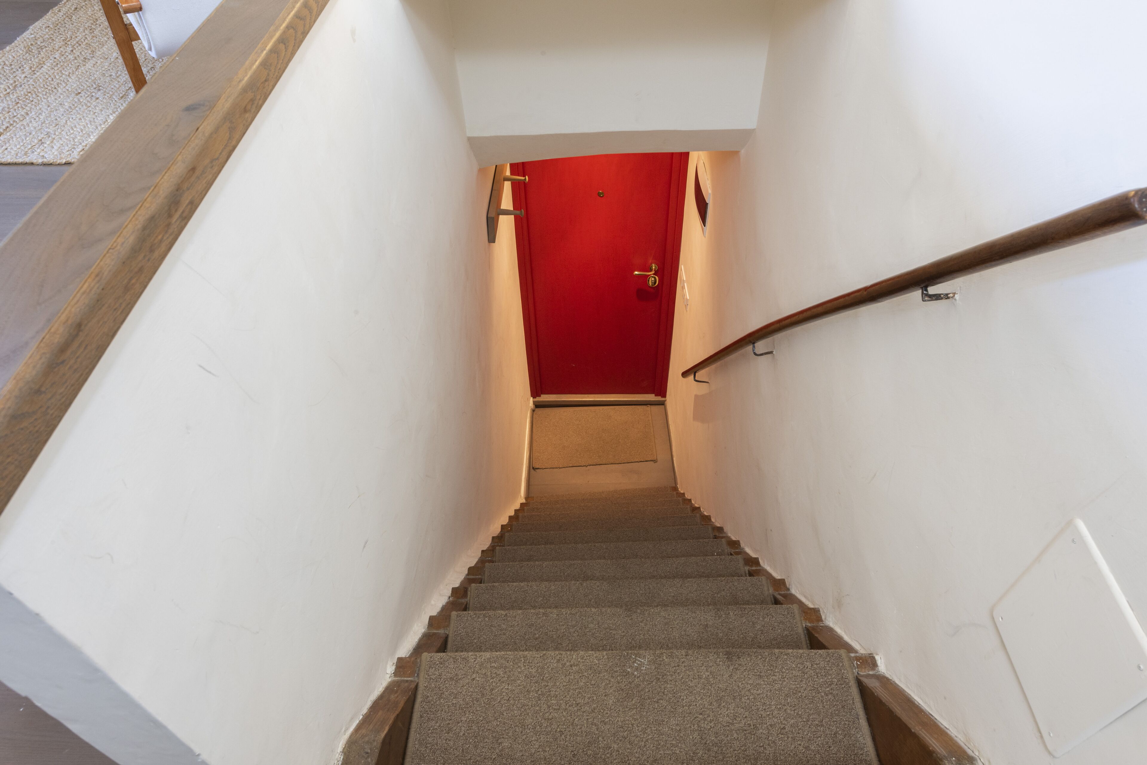 Una escalera con una puerta roja en la parte superior, paredes blancas y barandal de madera.