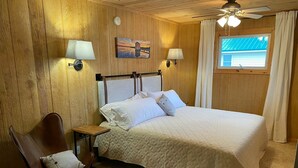 A bedroom with a bed, two wall-mounted lamps, a painting, a ceiling fan, and a window with curtains. - River Bend Cottage (White Cloud)