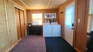 A narrow hallway with wooden walls and a carpeted floor. There are two white washing machines and a small table with a plant. - River Bend Cottage (White Cloud)
