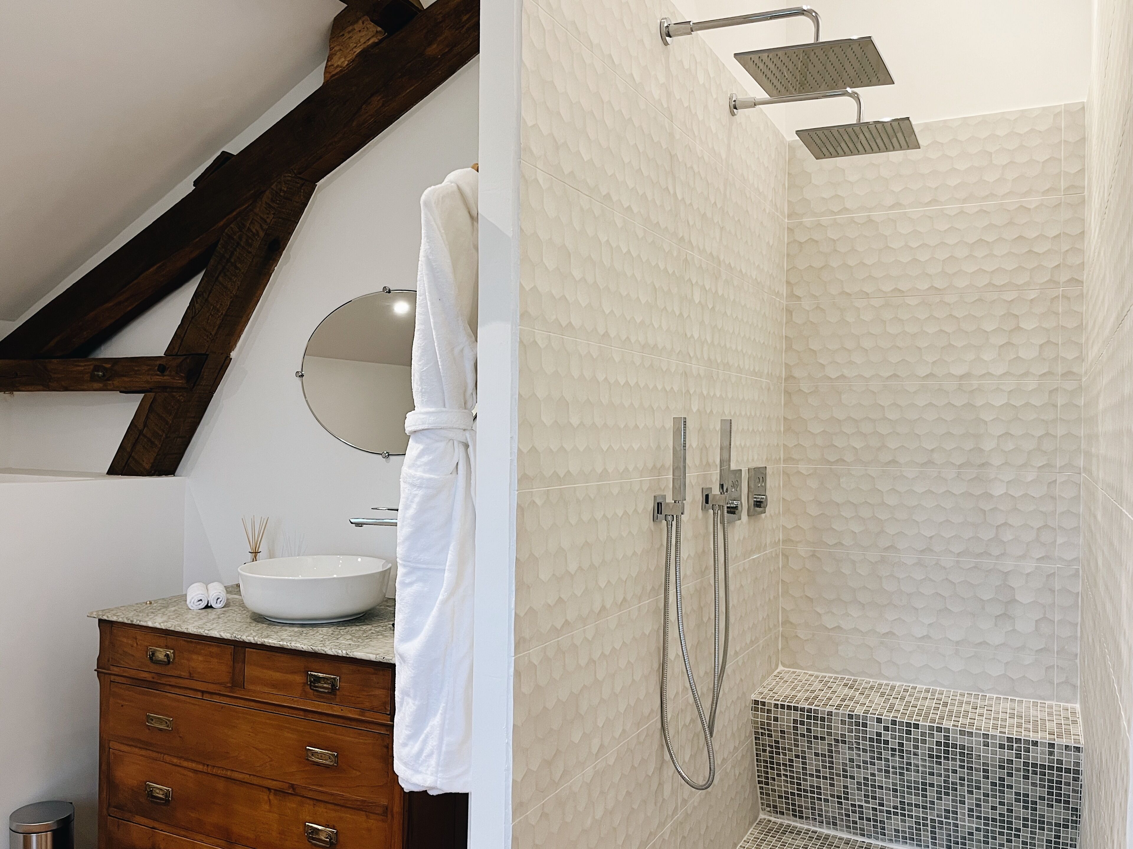 Ein Badezimmer mit Holzwaschtisch, einem runden Spiegel, einem weißen Waschbecken und einer Dusche mit strukturierter Wand.