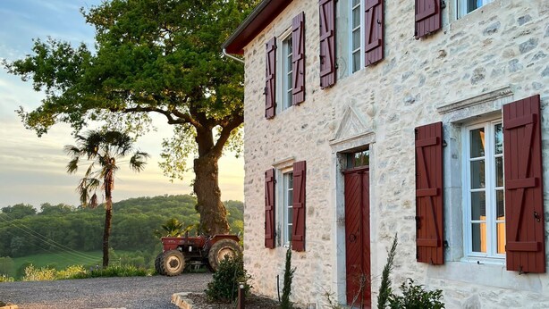Un edificio de piedra con contraventanas rojas, un camino de grava y un tractor aparcado cerca.