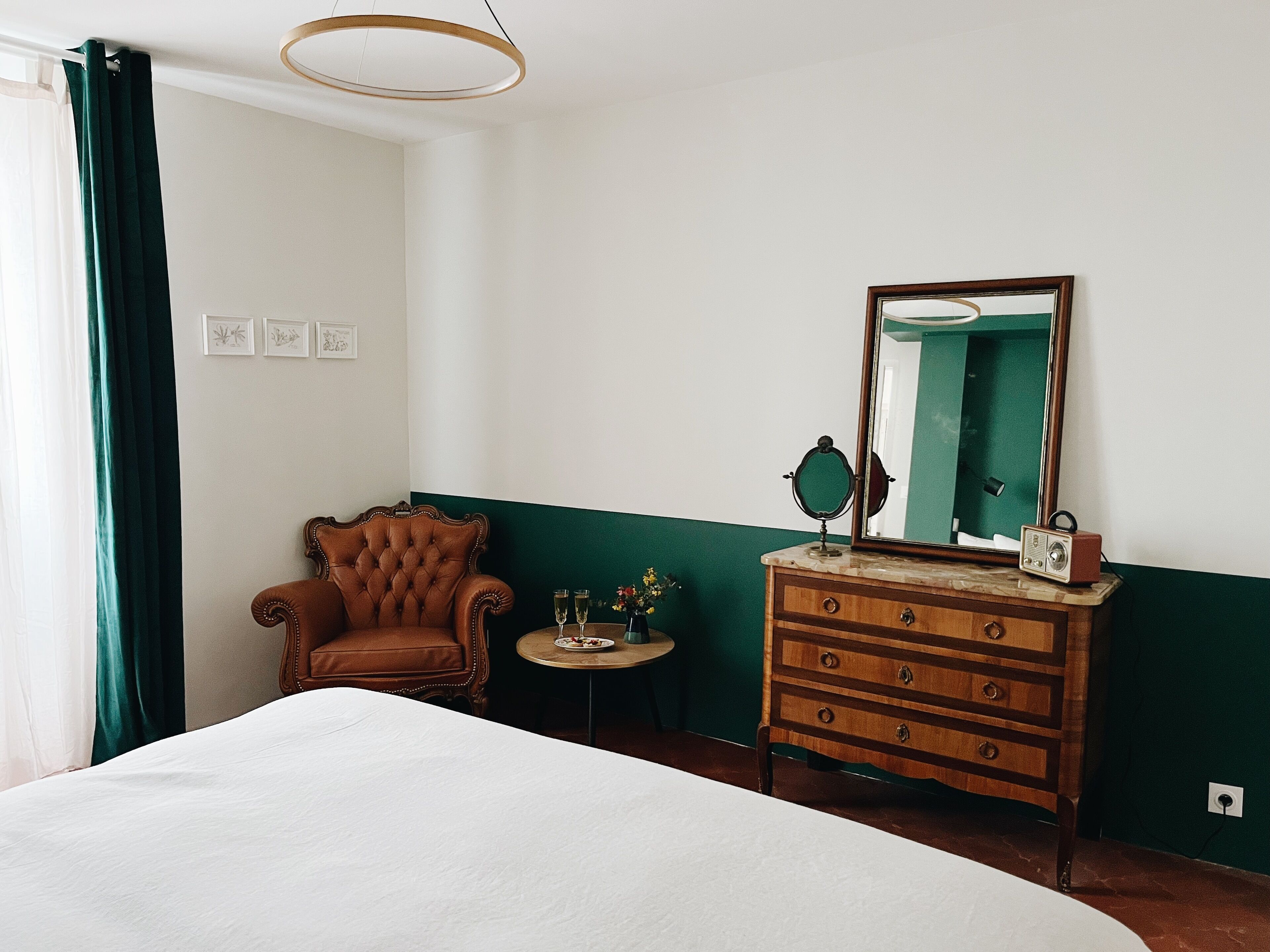 A hotel room with a bed, a wooden dresser, a chair, a round table with a mirror, and a green wall.