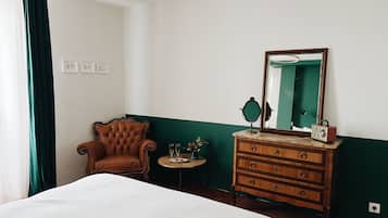 A hotel room with a bed, a wooden dresser, a chair, a round table with a mirror, and a green wall.