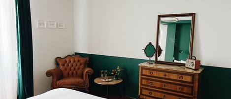 A hotel room with a bed, a wooden dresser, a chair, a round table with a mirror, and a green wall.